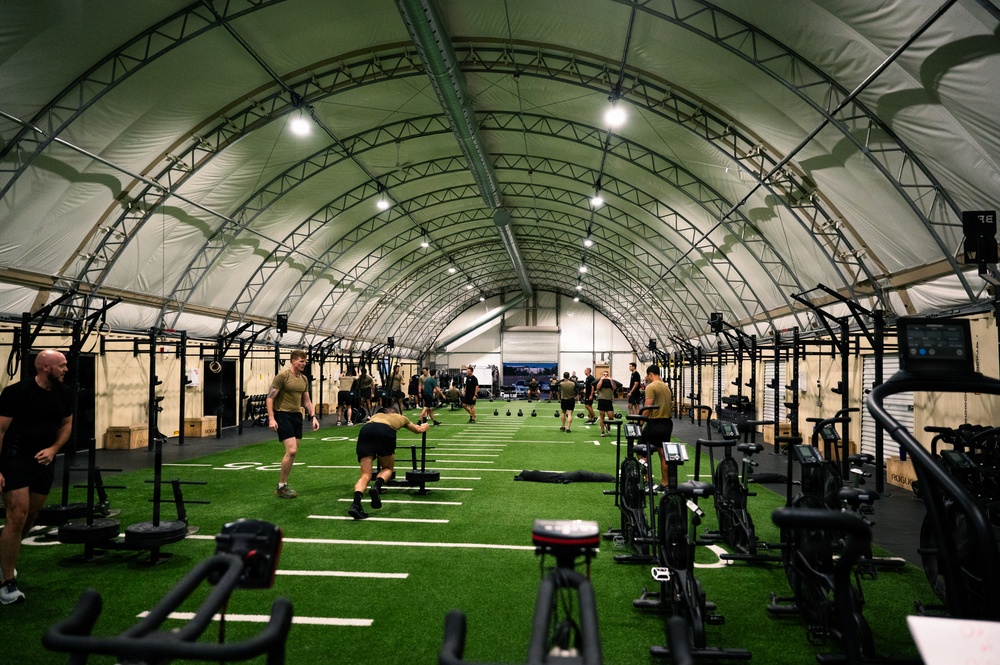 A large military fitness facility housed under an arched tension fabric structure with artificial turf flooring marked with yard lines. Multiple personnel in military-style physical training uniforms are engaged in various exercise activities across the expansive space. The facility is well-equipped with racks, stationary bikes, sleds, kettlebells, and other functional fitness equipment arranged along the sides and center.