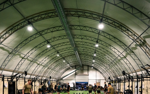 A large military fitness facility housed under an arched tension fabric structure with artificial turf flooring marked with yard lines. Multiple personnel in military-style physical training uniforms are engaged in various exercise activities across the expansive space. The facility is well-equipped with racks, stationary bikes, sleds, kettlebells, and other functional fitness equipment arranged along the sides and center.