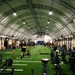 A large military fitness facility housed under an arched tension fabric structure with artificial turf flooring marked with yard lines. Multiple personnel in military-style physical training uniforms are engaged in various exercise activities across the expansive space. The facility is well-equipped with racks, stationary bikes, sleds, kettlebells, and other functional fitness equipment arranged along the sides and center.