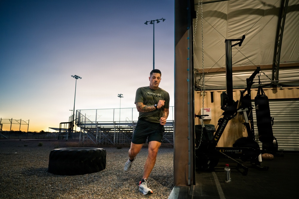 A tattooed male athlete is performing a running or high-knee exercise outdoors near the entrance of an open-air gym facility at dusk or dawn. The setting appears to be a military or athletic compound, with stadium bleachers, flood lights, and a large tractor tire visible in the background. The gym interior contains Concept2 rowing machines, a cable machine, and a punching bag.
