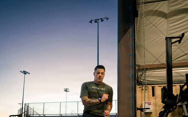 A tattooed male athlete is performing a running or high-knee exercise outdoors near the entrance of an open-air gym facility at dusk or dawn. The setting appears to be a military or athletic compound, with stadium bleachers, flood lights, and a large tractor tire visible in the background. The gym interior contains Concept2 rowing machines, a cable machine, and a punching bag.