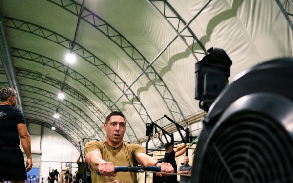A man in a tan/olive athletic shirt is using a Concept2 rowing machine inside a large indoor sports facility with an arched fabric roof structure. The facility appears to be hosting a fitness event or training session, with multiple participants and equipment visible in the background. The image is taken from a low angle close to the rowing machine, emphasizing the athlete's effort and the equipment.