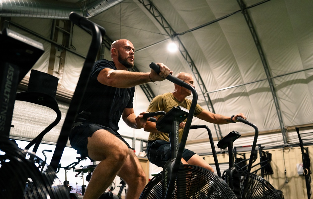 Two men are exercising on Assault Fitness air bikes inside what appears to be a military or temporary fabric-structure gym facility. The foreground subject is a bald, muscular man in a black t-shirt and shorts, pedaling intensely, while a second man in a yellow-green shirt rides behind him. The low-angle shot emphasizes the physical exertion and the industrial setting of the workout space.