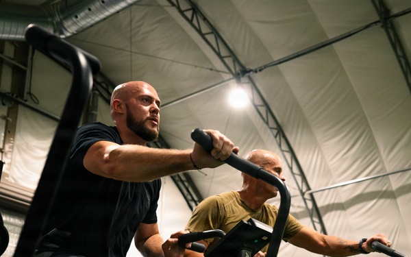 Two men are exercising on Assault Fitness air bikes inside what appears to be a military or temporary fabric-structure gym facility. The foreground subject is a bald, muscular man in a black t-shirt and shorts, pedaling intensely, while a second man in a yellow-green shirt rides behind him. The low-angle shot emphasizes the physical exertion and the industrial setting of the workout space.
