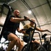 Two men are exercising on Assault Fitness air bikes inside what appears to be a military or temporary fabric-structure gym facility. The foreground subject is a bald, muscular man in a black t-shirt and shorts, pedaling intensely, while a second man in a yellow-green shirt rides behind him. The low-angle shot emphasizes the physical exertion and the industrial setting of the workout space.