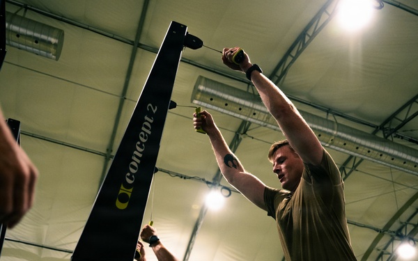 Two individuals are working with Concept2 rowing machines inside a large indoor facility with an arched ceiling structure. The person in the foreground is standing upright with arms raised, appearing to handle the rowing machine's chain or handle mechanism. The setting appears to be a military or institutional fitness facility based on the architecture and attire.