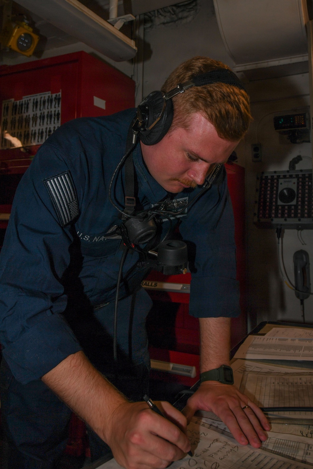 A U.S. Navy sailor in a blue coverall uniform is writing on documents or logs at a workstation inside what appears to be a shipboard workspace or maintenance area. He is wearing communication headphones around his neck and has a subdued American flag patch on his left shoulder. The environment suggests an operational or engineering space aboard a naval vessel.