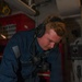 A U.S. Navy sailor in a blue coverall uniform is writing on documents or logs at a workstation inside what appears to be a shipboard workspace or maintenance area. He is wearing communication headphones around his neck and has a subdued American flag patch on his left shoulder. The environment suggests an operational or engineering space aboard a naval vessel.