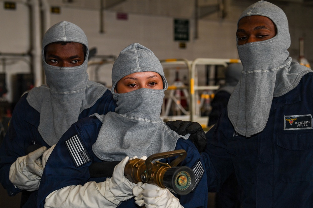 Three military personnel wearing gray fire-resistant flash hoods and dark blue coveralls are posed together, with the center individual holding a brass fire hose nozzle. They appear to be U.S. Navy sailors conducting or preparing for firefighting training, based on their uniforms and equipment. The setting appears to be an indoor hangar or ship bay environment.