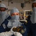Three military personnel wearing gray fire-resistant flash hoods and dark blue coveralls are posed together, with the center individual holding a brass fire hose nozzle. They appear to be U.S. Navy sailors conducting or preparing for firefighting training, based on their uniforms and equipment. The setting appears to be an indoor hangar or ship bay environment.