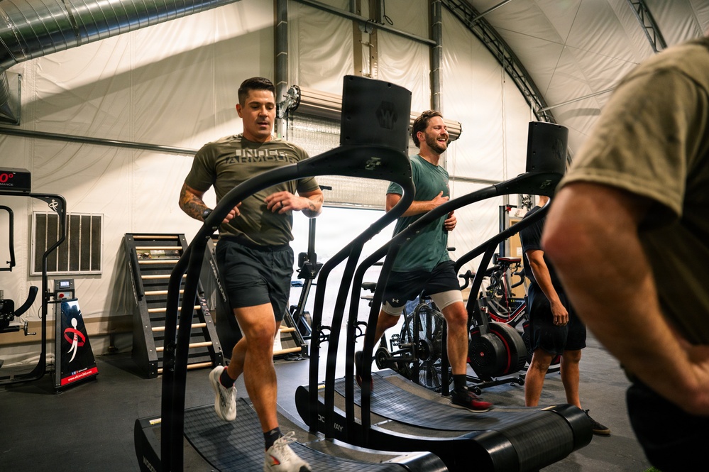 Three men are exercising on curved treadmills inside a well-equipped gym facility with a tent-like or fabric-walled structure. The men appear to be engaged in an energetic cardio workout, with two clearly visible individuals wearing athletic clothing including Under Armour apparel. The gym contains various fitness equipment including air bikes, plyometric boxes, and other training apparatus.