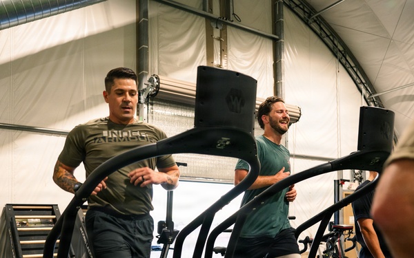 Three men are exercising on curved treadmills inside a well-equipped gym facility with a tent-like or fabric-walled structure. The men appear to be engaged in an energetic cardio workout, with two clearly visible individuals wearing athletic clothing including Under Armour apparel. The gym contains various fitness equipment including air bikes, plyometric boxes, and other training apparatus.
