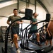 Three men are exercising on curved treadmills inside a well-equipped gym facility with a tent-like or fabric-walled structure. The men appear to be engaged in an energetic cardio workout, with two clearly visible individuals wearing athletic clothing including Under Armour apparel. The gym contains various fitness equipment including air bikes, plyometric boxes, and other training apparatus.