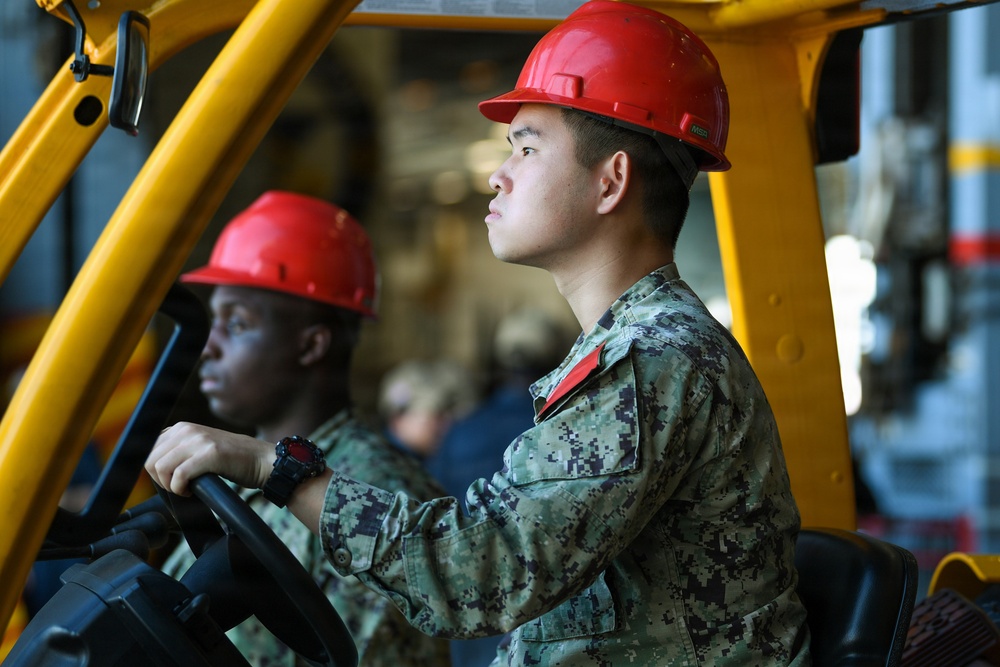 Two military personnel wearing red hard hats and digital camouflage uniforms (consistent with U.S. Navy NWU Type III pattern) are operating or riding in a yellow forklift or industrial vehicle. The foreground subject is at the steering wheel, looking forward with focused attention. The setting appears to be an industrial or shipboard environment.