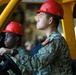 Two military personnel wearing red hard hats and digital camouflage uniforms (consistent with U.S. Navy NWU Type III pattern) are operating or riding in a yellow forklift or industrial vehicle. The foreground subject is at the steering wheel, looking forward with focused attention. The setting appears to be an industrial or shipboard environment.