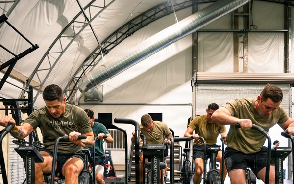 A group of individuals wearing matching olive/tan military-style t-shirts and dark shorts are intensely working out on Assault Fitness air bikes in what appears to be a military or tactical fitness facility. The gym is housed under an arched fabric/tensile structure with artificial turf flooring and professional fitness equipment. A digital timer on the left wall displays '1:00:00', suggesting a timed workout session is in progress.