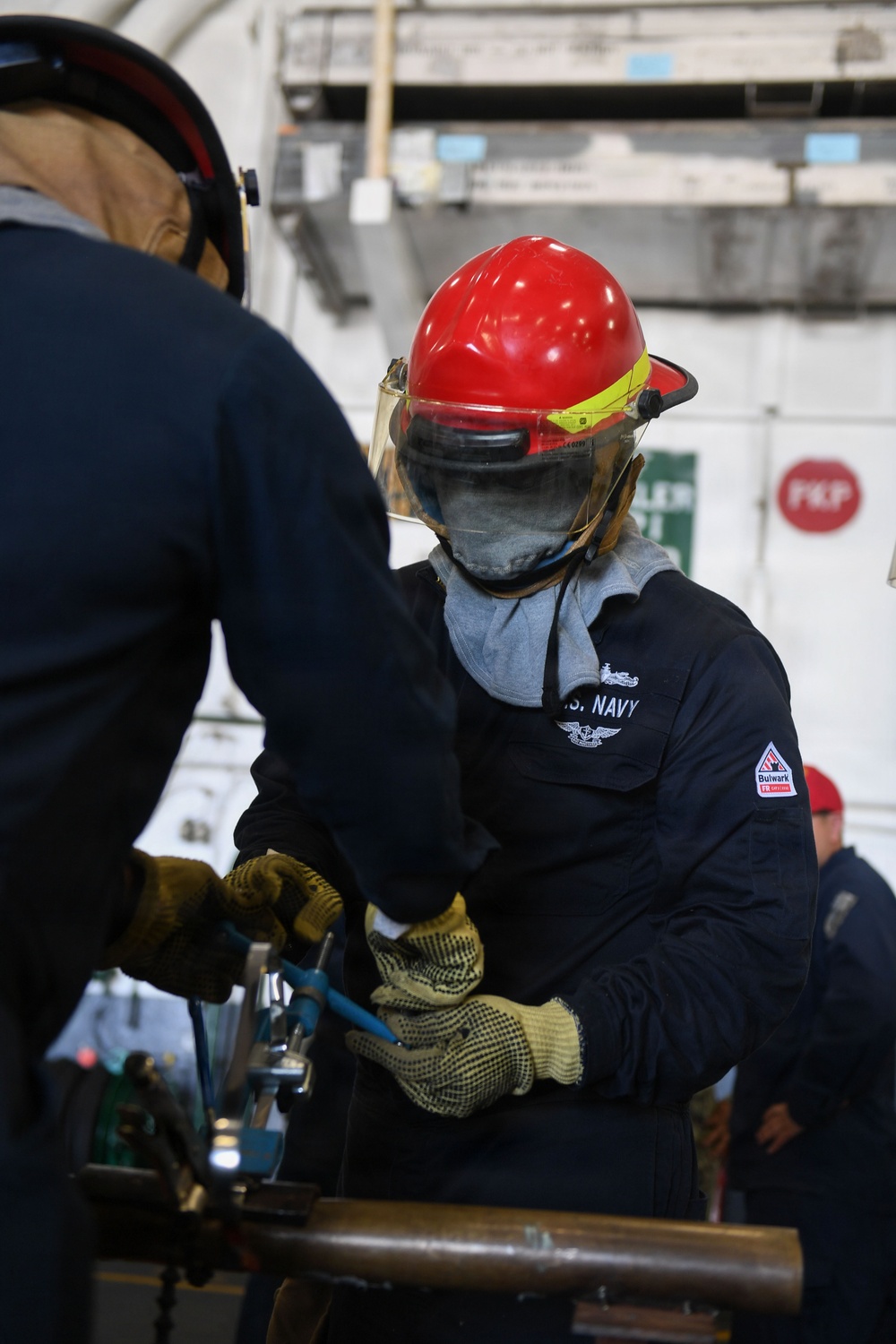 A U.S. Navy sailor wearing a red hard hat with face shield, flame-resistant Bulwark coveralls, and work gloves is engaged in a hands-on technical task alongside another person. The setting appears to be aboard a naval vessel, indicated by the industrial interior environment and Navy-branded gear. A third person in a red cap is visible in the background.