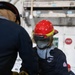 A U.S. Navy sailor wearing a red hard hat with face shield, flame-resistant Bulwark coveralls, and work gloves is engaged in a hands-on technical task alongside another person. The setting appears to be aboard a naval vessel, indicated by the industrial interior environment and Navy-branded gear. A third person in a red cap is visible in the background.