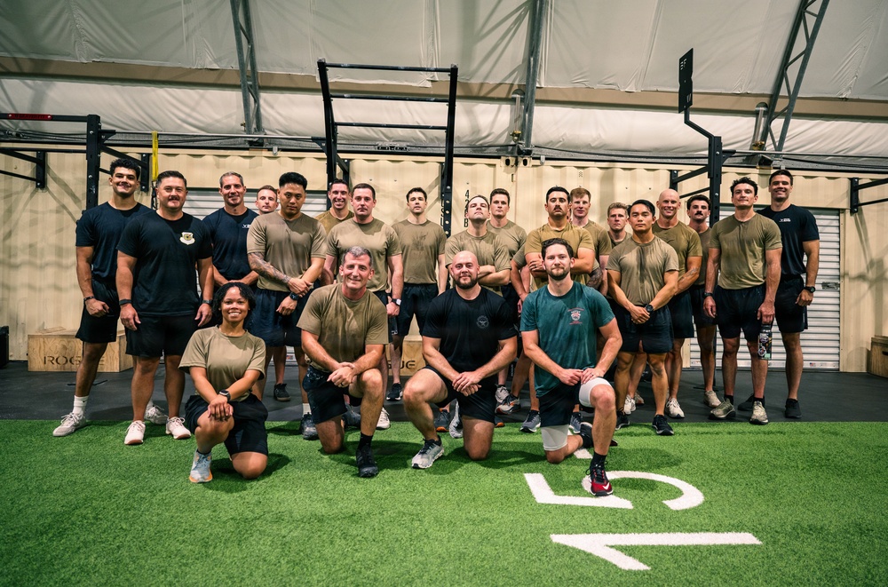 A large group of approximately 25 individuals poses for a group photo inside what appears to be a military or tactical fitness facility. Most participants are wearing olive/tan military-style t-shirts and dark shorts, suggesting a uniformed unit or training group. The setting is an indoor gym with turf flooring, pull-up rigs, and plywood box equipment.
