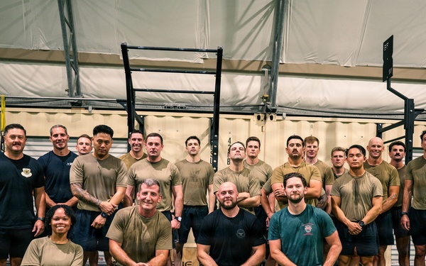 A large group of approximately 25 individuals poses for a group photo inside what appears to be a military or tactical fitness facility. Most participants are wearing olive/tan military-style t-shirts and dark shorts, suggesting a uniformed unit or training group. The setting is an indoor gym with turf flooring, pull-up rigs, and plywood box equipment.