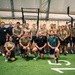 A large group of approximately 25 individuals poses for a group photo inside what appears to be a military or tactical fitness facility. Most participants are wearing olive/tan military-style t-shirts and dark shorts, suggesting a uniformed unit or training group. The setting is an indoor gym with turf flooring, pull-up rigs, and plywood box equipment.