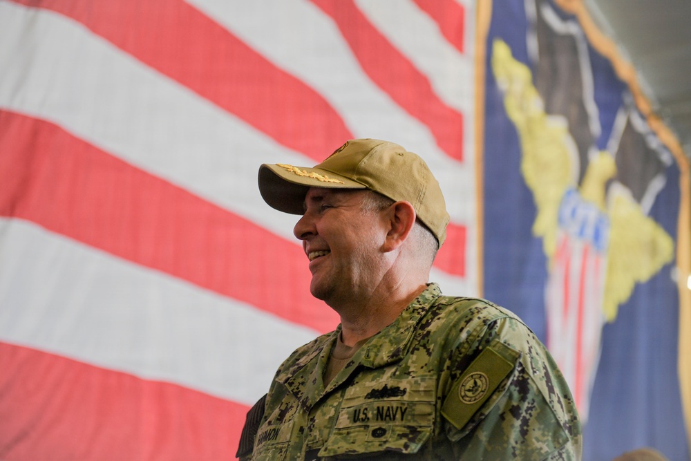 A U.S. Navy servicemember in digital camouflage uniform is smiling, photographed in front of a large American flag and what appears to be a military unit or command flag. The individual is wearing a tan/khaki baseball cap with what appears to be a command insignia, and their uniform name tape reads 'U.S. NAVY'. The setting appears to be an indoor military ceremony or event.