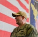 A U.S. Navy servicemember in digital camouflage uniform is smiling, photographed in front of a large American flag and what appears to be a military unit or command flag. The individual is wearing a tan/khaki baseball cap with what appears to be a command insignia, and their uniform name tape reads 'U.S. NAVY'. The setting appears to be an indoor military ceremony or event.