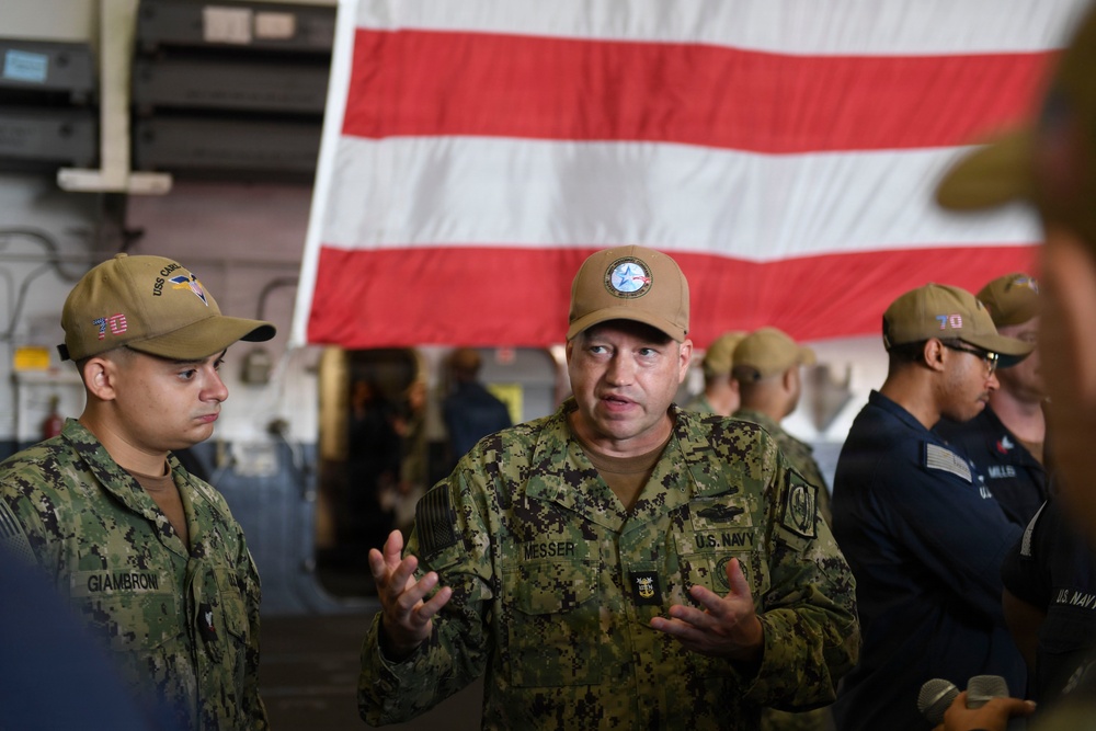 Two U.S. Navy personnel are engaged in conversation aboard what appears to be a naval vessel, likely in a hangar bay. The central figure, identified by name tape as 'MESSER,' is speaking animatedly with hands gesturing, while 'GIAMBRONI' listens attentively. A large American flag is displayed prominently in the background.