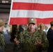 Two U.S. Navy personnel are engaged in conversation aboard what appears to be a naval vessel, likely in a hangar bay. The central figure, identified by name tape as 'MESSER,' is speaking animatedly with hands gesturing, while 'GIAMBRONI' listens attentively. A large American flag is displayed prominently in the background.