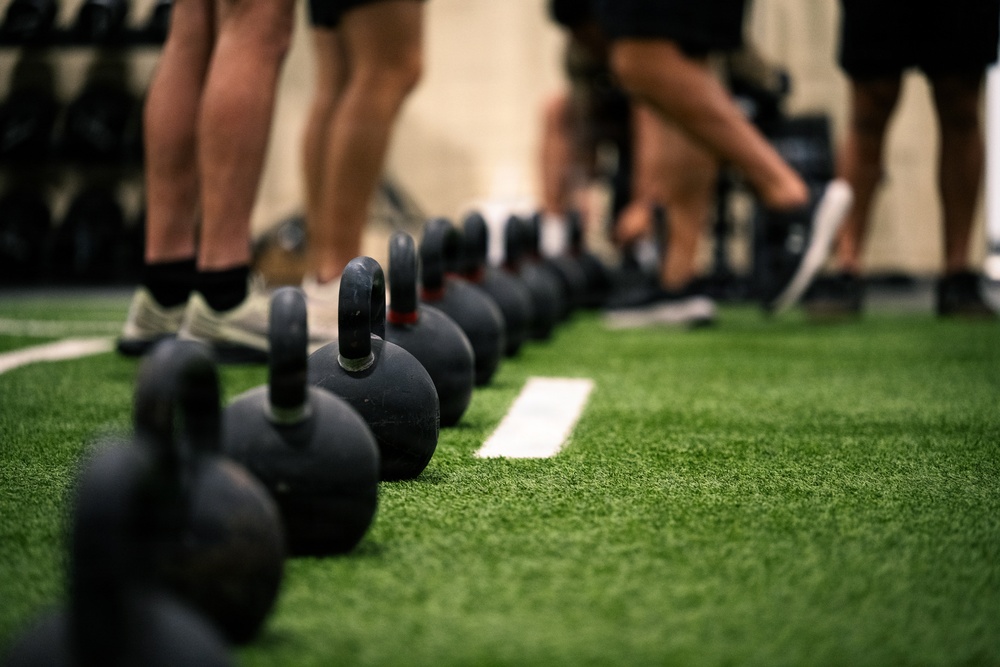 A row of black cast-iron kettlebells is lined up on an artificial green turf floor in what appears to be a fitness facility or gym. Multiple people are visible in the background, identifiable only by their legs and feet, suggesting a group training session is underway. The foreground kettlebells are in sharp focus while the background elements are intentionally blurred using shallow depth of field.
