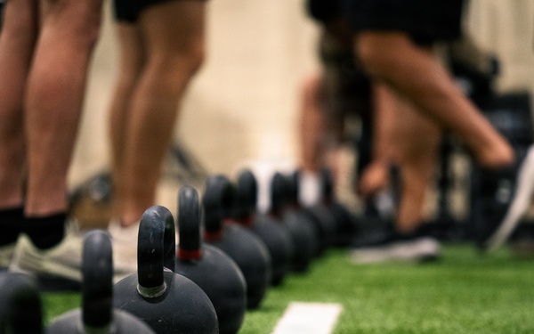 A row of black cast-iron kettlebells is lined up on an artificial green turf floor in what appears to be a fitness facility or gym. Multiple people are visible in the background, identifiable only by their legs and feet, suggesting a group training session is underway. The foreground kettlebells are in sharp focus while the background elements are intentionally blurred using shallow depth of field.