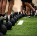 A row of black cast-iron kettlebells is lined up on an artificial green turf floor in what appears to be a fitness facility or gym. Multiple people are visible in the background, identifiable only by their legs and feet, suggesting a group training session is underway. The foreground kettlebells are in sharp focus while the background elements are intentionally blurred using shallow depth of field.