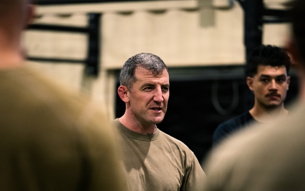 A middle-aged man with gray-streaked hair is speaking to a group of individuals in what appears to be a military or fitness facility. He is wearing an olive/tan t-shirt, consistent with military physical training attire, and appears to be addressing or briefing the group. Several other individuals in similar attire are visible in the foreground and background, slightly out of focus.