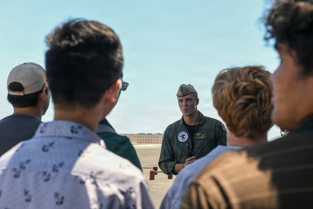 A military aviator in a green flight suit and garrison cap addresses a small group of civilians on what appears to be an airfield tarmac. The setting suggests an outreach event, tour, or recruitment briefing at a military air installation. The speaker is gesturing and appears to be actively explaining something to the assembled group.