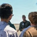 A military aviator in a green flight suit and garrison cap addresses a small group of civilians on what appears to be an airfield tarmac. The setting suggests an outreach event, tour, or recruitment briefing at a military air installation. The speaker is gesturing and appears to be actively explaining something to the assembled group.