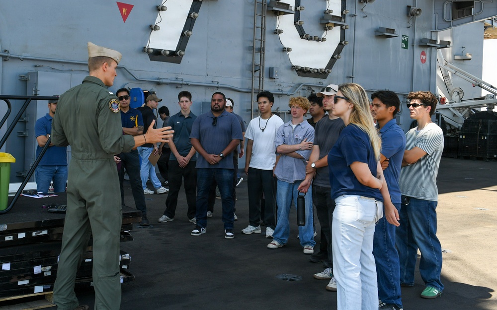 A military officer in olive green flight suit and white cap is briefing a group of approximately 12-15 civilians on the deck of a naval vessel. The large hull number '79' is partially visible on the ship's superstructure in the background, suggesting this is likely USS Ronald Reagan (CVN-79) or a similar numbered vessel. The scene appears to be an organized tour or recruitment/outreach event.