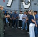 A military officer in olive green flight suit and white cap is briefing a group of approximately 12-15 civilians on the deck of a naval vessel. The large hull number '79' is partially visible on the ship's superstructure in the background, suggesting this is likely USS Ronald Reagan (CVN-79) or a similar numbered vessel. The scene appears to be an organized tour or recruitment/outreach event.
