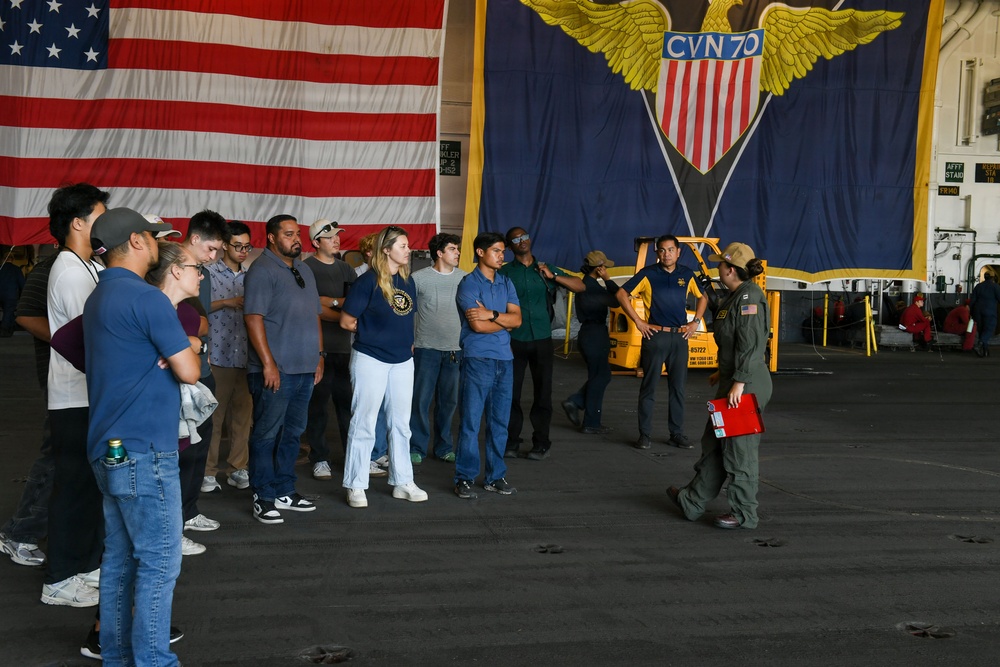 A group of civilians is receiving a tour or briefing aboard USS Carl Vinson (CVN-70), a U.S. Navy aircraft carrier, in the hangar bay. A military personnel member in flight suit is addressing the group while holding a red clipboard or binder. Large American flag and the ship's official CVN-70 insignia banner are prominently displayed in the background.