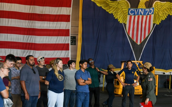 A group of civilians is receiving a tour or briefing aboard USS Carl Vinson (CVN-70), a U.S. Navy aircraft carrier, in the hangar bay. A military personnel member in flight suit is addressing the group while holding a red clipboard or binder. Large American flag and the ship's official CVN-70 insignia banner are prominently displayed in the background.