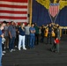 A group of civilians is receiving a tour or briefing aboard USS Carl Vinson (CVN-70), a U.S. Navy aircraft carrier, in the hangar bay. A military personnel member in flight suit is addressing the group while holding a red clipboard or binder. Large American flag and the ship's official CVN-70 insignia banner are prominently displayed in the background.