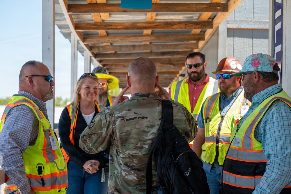 A military personnel in camouflage uniform is briefing or conversing with a group of civilian construction or safety workers wearing high-visibility vests. The meeting appears to take place under a partially constructed or damaged covered walkway at what looks like a construction or disaster recovery site. The interaction suggests a coordination meeting between military and civilian project personnel.