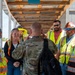 A military personnel in camouflage uniform is briefing or conversing with a group of civilian construction or safety workers wearing high-visibility vests. The meeting appears to take place under a partially constructed or damaged covered walkway at what looks like a construction or disaster recovery site. The interaction suggests a coordination meeting between military and civilian project personnel.