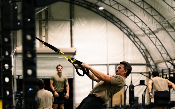 A group of individuals in matching olive/tan athletic shirts and dark shorts are training inside a large indoor facility with an arched, fabric-covered roof. The central figure is performing a TRX suspension trainer row exercise, leaning back at an angle while gripping the straps. Multiple participants are engaged in various functional fitness exercises across a turf floor marked with white yard lines.