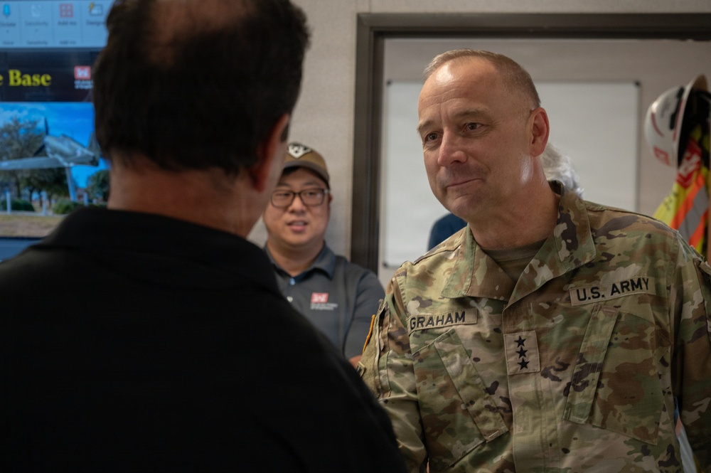 A U.S. Army Major General named Graham, identifiable by two stars on his uniform and name tape, is engaged in conversation with a civilian whose back is to the camera. The setting appears to be an indoor facility, possibly a military installation or operations center. A second civilian in a gray polo shirt and cap observes the conversation in the background.