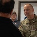 A U.S. Army Major General named Graham, identifiable by two stars on his uniform and name tape, is engaged in conversation with a civilian whose back is to the camera. The setting appears to be an indoor facility, possibly a military installation or operations center. A second civilian in a gray polo shirt and cap observes the conversation in the background.