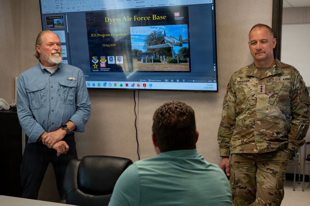 A briefing or presentation is taking place in a conference room at or related to Dyess Air Force Base. A U.S. Army Lieutenant General (three stars) named Graham stands to the right in military camouflage uniform, while a civilian in a blue shirt stands to the left. A seated individual with their back to the camera appears to be the audience, with a large display screen showing a 'Dyess Air Force Base - B21 Program Overview' presentation dated 18 Aug 2026.