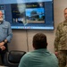 A briefing or presentation is taking place in a conference room at or related to Dyess Air Force Base. A U.S. Army Lieutenant General (three stars) named Graham stands to the right in military camouflage uniform, while a civilian in a blue shirt stands to the left. A seated individual with their back to the camera appears to be the audience, with a large display screen showing a 'Dyess Air Force Base - B21 Program Overview' presentation dated 18 Aug 2026.