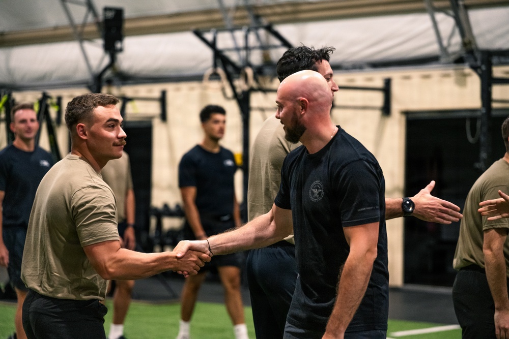 Two men are shaking hands in what appears to be a military or law enforcement fitness facility. The individuals are wearing a mix of olive/tan and black athletic t-shirts, consistent with military physical training attire. Several other similarly dressed individuals are visible in the background of the indoor gym setting.