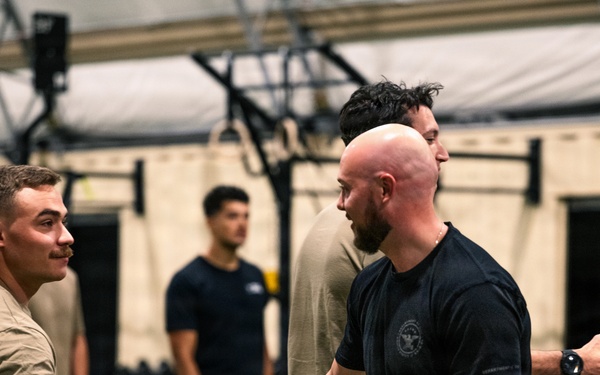 Two men are shaking hands in what appears to be a military or law enforcement fitness facility. The individuals are wearing a mix of olive/tan and black athletic t-shirts, consistent with military physical training attire. Several other similarly dressed individuals are visible in the background of the indoor gym setting.