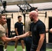 Two men are shaking hands in what appears to be a military or law enforcement fitness facility. The individuals are wearing a mix of olive/tan and black athletic t-shirts, consistent with military physical training attire. Several other similarly dressed individuals are visible in the background of the indoor gym setting.